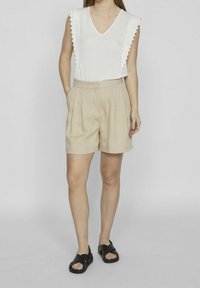 Bílý top s výstřihem do V a zvlněnými rukávy, v kombinaci s béžovými skládacími šortkami. Modelka má na sobě černé sandály s křížovými řemínky. Hladký povrch materiálu.