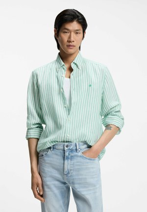 Giovane uomo con capelli neri che indossa una camicia a righe verdi e bianche con bottoni sopra una canottiera bianca e jeans azzurri, con una mano in tasca.