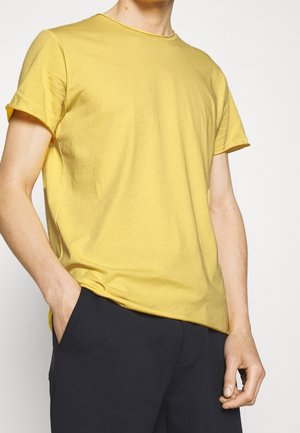 Mand iført en ensfarvet gul kortærmet t-shirt med den ene hånd i lommen på sorte bukser, stående mod en hvid baggrund.