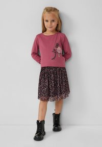 Junges Mädchen mit langen blonden Haaren, das ein rosa Langarmoberteil mit Katzen- und Ballerina-Design, einen Blumenrock und schwarze Schnürstiefel trägt.