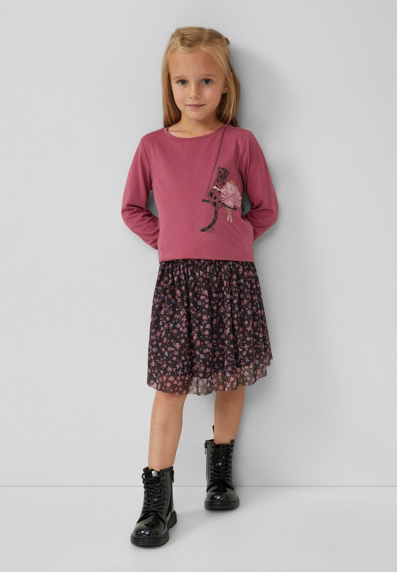 Junges Mädchen mit langen blonden Haaren, das ein rosa Langarmoberteil mit Katzen- und Ballerina-Design, einen Blumenrock und schwarze Schnürstiefel trägt.