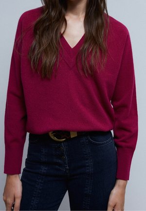 Woman wearing a dark pink V-neck sweater tucked into high-waisted dark blue jeans with a black belt featuring a decorative buckle.