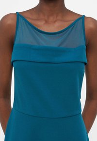 Μπλε-πράσινο φόρεμα χωρίς μανίκια με διαφανές πλεκτό μέρος στο πάνω μέρος και διπλωμένες λεπτομέρειες υφάσματος στο στήθος, φορεμένο από ένα άτομο με σκούρο χρώμα δέρματος.