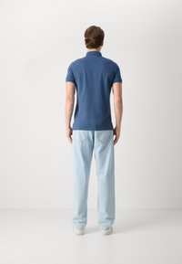 Homme debout de dos, portant un polo bleu à manches courtes, un jean bleu clair et des baskets blanches sur un fond uni.