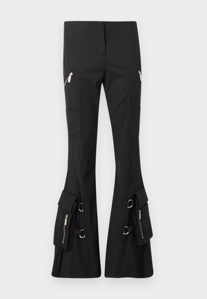 Junge Frau trägt ein weißes T-Shirt mit Globus-Design, schwarze Cargohosen mit Taschen, schwarze Schuhe und eine schwarze Baseballkappe.