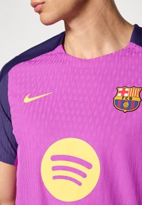 Nogometna majica Barcelona z vijolično telo, temno modrima rokavoma, teksturirano tkanino, velikim rumenim logotipom Spotify in grbom kluba.