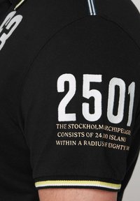 Black polo shirt featuring large white numeral "2501" and text in gold. Accents include yellow and white stripes on the collar.