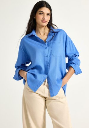 Kvinde iført en løs, knap-til skjorte i klar blå med ærmerne rullet op og beige bukser, stående med hænderne i lommerne mod en hvid baggrund.