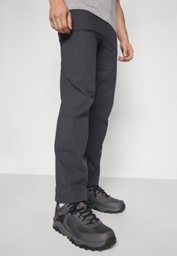 Mann iført mørkegrå bukser og grå og svarte tursko, stående på et lyst gulv mot en ensfarget vegg.