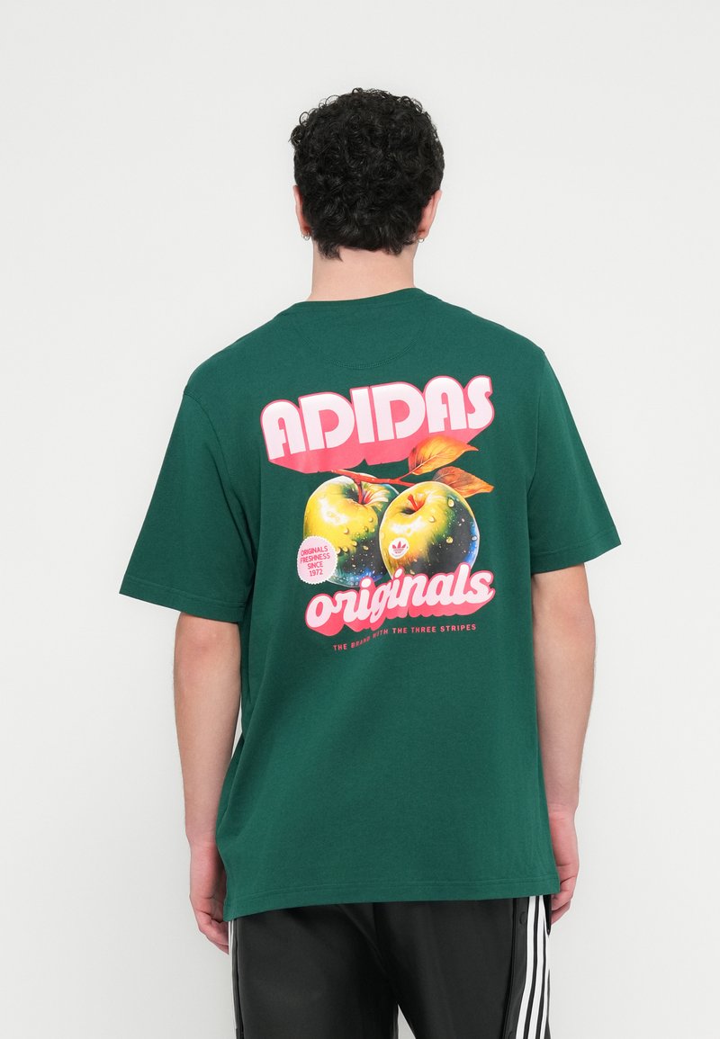 Zaļš kokvilnas T-krekls ar krāsainu ābolu grafiku aizmugurē, uz kura rakstīts "ADIDAS" un "originals" ar treknu rozā un baltu tipogrāfiju.