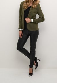 Olivgrön kavaj med vita randiga manschetter, matchad med svarta imiterade läderbyxor och svarta högklackade skor. Slät textur och åtsittande design.