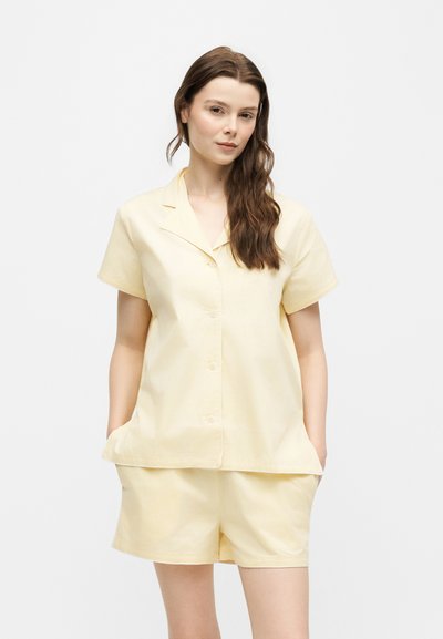 Jeune femme aux longs cheveux bruns portant une chemise boutonnée à manches courtes jaune pâle et un short assorti, debout, mains dans les poches.