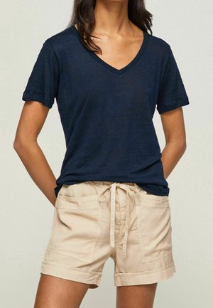 Femme portant un t-shirt à manches courtes bleu marine à col en V et un short beige avec cordon de serrage et poches avant, debout les mains dans le dos.