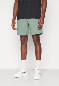 Shorts vert menthe à coupe droite, arborant un logo noir "BOSS". Portés avec des chaussettes blanches et des baskets, associés à un t-shirt noir.