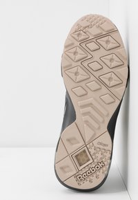 Semelle de chaussure Reebok, présentant un caoutchouc beige texturé avec des motifs en forme de losange, un bord supérieur noir, et une section de coussin hexagonale visible.