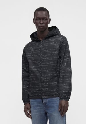Ung mand iført en sort lynlåshættetrøje med gentaget "EMPORIO ARMANI" tekst og blå jeans, stående mod en ensartet lys baggrund.