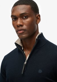 Homme à la peau foncée portant un pull à demi-fermeture éclair bleu marine par-dessus une chemise beige à col, regardant directement la caméra avec une expression neutre.