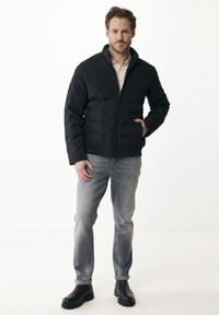 Muž stojí s rukou v kapse, oblečený v černé péřové bundě, béžovém polo tričku, šedých džínách a černých botách na bílém pozadí.