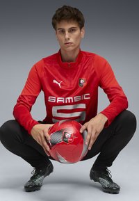 Maglia sportiva rossa con design testurizzato, pantaloni neri e scarpette nere. Il giocatore tiene in mano un pallone da calcio rosso e nero con il logo del club.