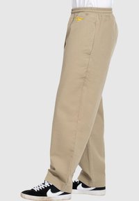 Beige Jogginghosen aus Baumwolle mit einem Kordelzug, seitlichen Taschen und einem kleinen gelben Logo in Hüfthöhe, kombiniert mit schwarzen Sneakern.