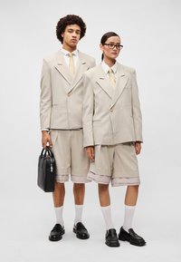 Blazers bege ajustados com lapelas pontudas, combinados com calções a condizer com bainhas ligeiramente onduladas. Bolsa preta e mocassins, meias brancas.