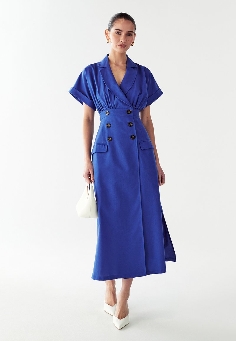 Karališkai mėlyna suknelė su dvigubos sagos dizainu, plačiomis trumpomis rankovėmis, šonine iškirpte ir priekiniame kišenėje, derinama su balta rankine ir aukštakulniais.