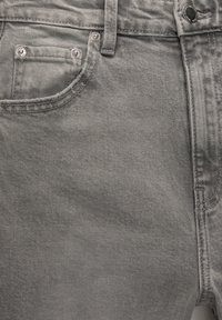 Gros plan sur un jean en denim gris montrant la poche avant, la poche à monnaie, la passants de ceinture, ainsi que les rivets et le bouton en métal argenté.
