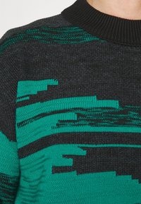Pull en maille noire avec des motifs abstraits en bleu sarcelle et vert foncé, présentant un col côtelé et un tricot texturé varié sur toute la surface.