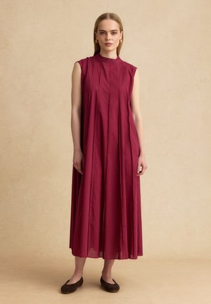 Mujer de pie con un vestido sin mangas, holgado, hasta el tobillo, de color burdeos con pliegues verticales y zapatos planos oscuros.