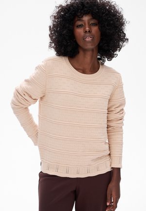 Vrouw met krullend haar die een gestructureerde beige trui en donkerbruine broek draagt, staande tegen een eenvoudige witte achtergrond.