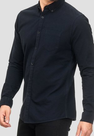 Mężczyzna ubrany w dopasowaną czarną koszulę na guziki z długim rękawem i kieszenią na piersi, stojący na tle jednolitego jasnoszarego tła.