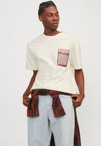 T-shirt couleur crème à manches courtes, avec un imprimé graphique de poche bordeaux, porté avec un jean bleu clair et une chemise à carreaux rouge nouée à la taille.