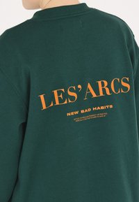 Mörkgrön sweatshirt med orange text på baksidan som läser "LES'ARCS" och "NEW BAD HABITS" med ytterligare definitioner nedan.
