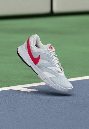 Scarpa da tennis Nike in grigio chiaro con il logo Swoosh rosso. Presenta una tomaia testurizzata, suola bianca e perforazioni per la ventilazione. Fluttua sopra la superficie del campo.