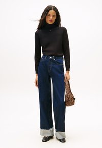 Donna con lunghi capelli ricci che indossa un dolcevita nero, jeans blu scuro a gamba larga risvoltati all'orlo, scarpe nere con borchie, mentre tiene una borsa con stampa leopardata.