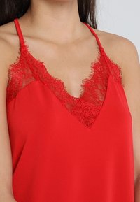 Rode mouwloze top met een V-hals afgezet met kant, gladde stof en fijne ruches aan het kant, wat een gelaagde uitstraling geeft.