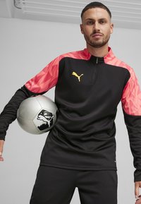 Schwarzes Sportoberteil mit Ärmeln im pinken geometrischen Muster, ein Modell steht und hält einen silbernen Fußball, präsentiert ein goldenes Puma-Logo und hat einen Reißverschlusskragen.