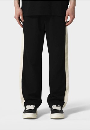 Sorte bukser med kremfargede side-striper, laget av et mykt stoff. Har en løs passform og elastisk liv. Brukt med svarte joggesko.