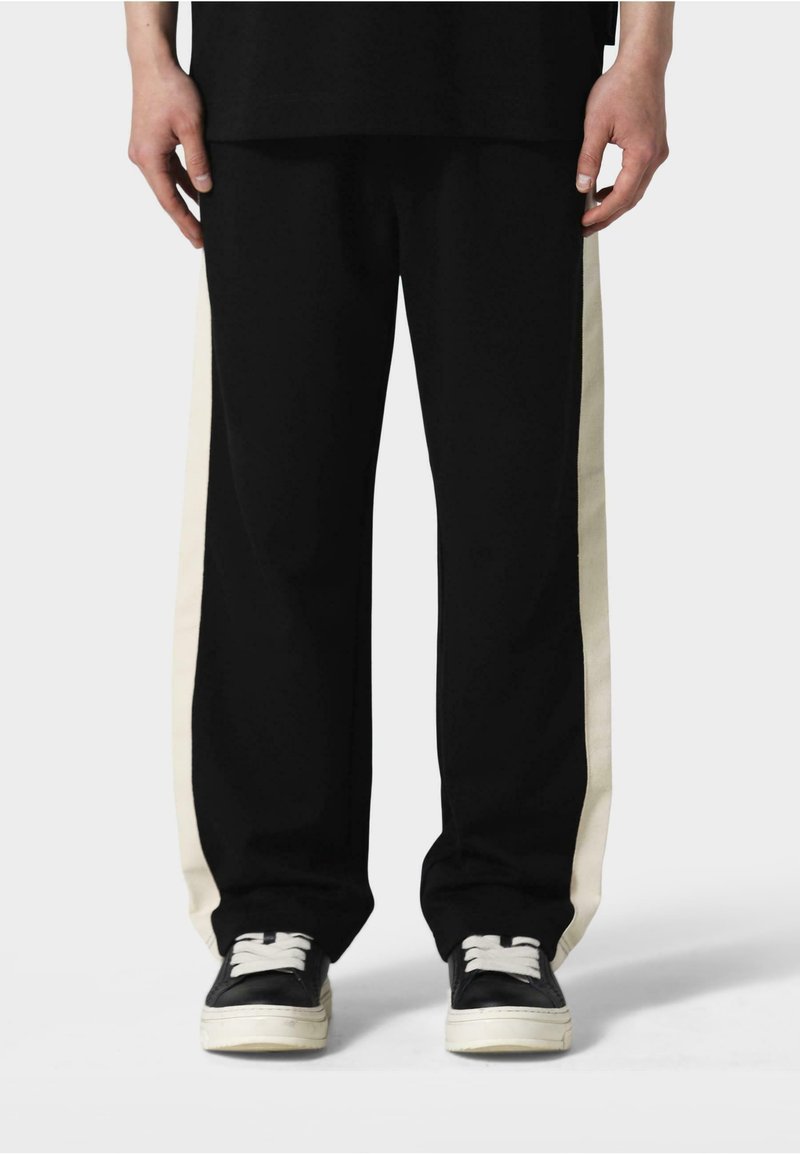 Sorte bukser med cremede sidestriber, lavet af et blødt stof. Har en løs pasform og elastisk talje. Båret med sorte sneakers.