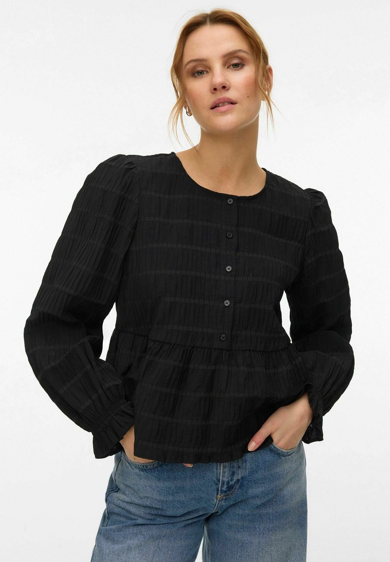 Femme à la peau claire et aux cheveux blonds portant un chemisier noir texturé à boutons avec des manches longues bouffantes et un jean bleu, les mains dans les poches.