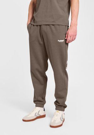 Mężczyzna ubrany w brązowe spodnie dresowe Hummel i białe trampki z szarymi wzorami w zygzak, stoi z rękami w kieszeniach.