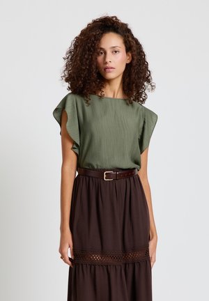 Frau mit lockigem Haar, die ein grünes, strukturiertes ärmelloses Oberteil und einen hoch taillierten dunkelbraunen Rock mit Spitzen-Details und Gürtel trägt.