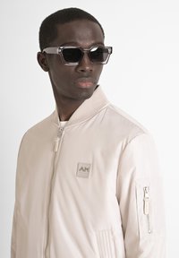 Chaqueta bomber beige con cremallera en la parte delantera y cuello de canalé; presenta un parche con logotipo y un bolsillo lateral. Gafas de sol negras con un marco grueso.