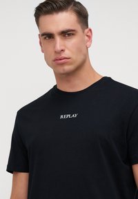 Camiseta de algodón negra con cuello redondo y mangas cortas, con un logo bordado en blanco que dice "REPLAY" en el pecho.
