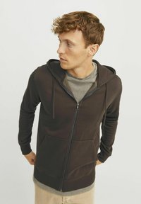 Sudadera con cremallera de color marrón oscuro, con un bolsillo tipo canguro, capucha ajustable con cordón y textura de tela suave. Camiseta gris visible debajo.