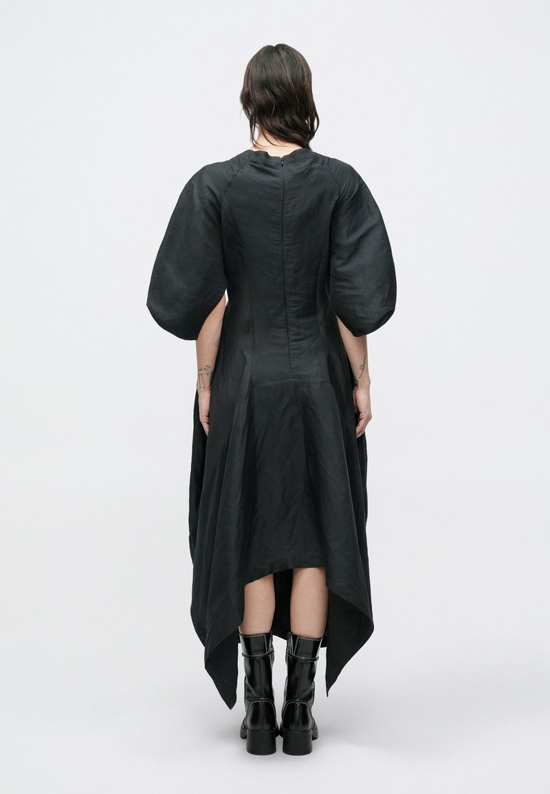 Vestito nero con maniche a sbuffo, orlo asimmetrico con parte posteriore più lunga, tessuto texturizzato e cerniera nascosta sul retro. Indossato con stivali neri alti fino al ginocchio.