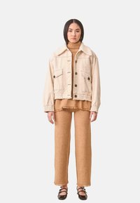 Chaqueta de mezclilla beige con cuello de piel de oveja, frente de botones y dos bolsillos en el pecho, combinada con una blusa marrón con flecos y pantalones de pierna ancha a juego.