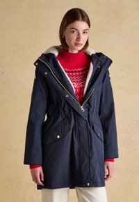 Donna che indossa un parka blu navy con cappuccio foderato in pelliccia, sopra un maglione rosso a fantasia e pantaloni bianchi, in piedi davanti a uno sfondo beige.
