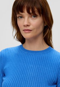 Blauer gerippter Strickpullover mit Rundhalsausschnitt. Die Textur ist glatt mit vertikalen Streifen. Das Model hat schulterlanges braunes Haar.
