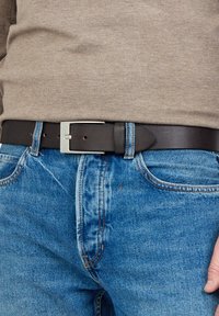 Mustat nahkavyöt, joissa on hopeinen metallisolki, käytetään sinisten farkkujen päällä, jotka ovat näkyvällä ompelulla ja sileällä pinnalla.